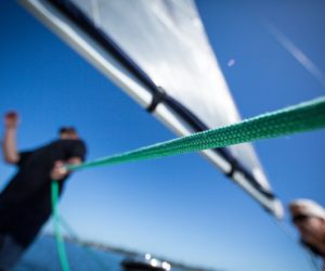 Taut green rope crosses a blurred low-angle view of two people and a sail