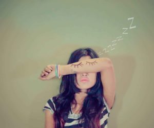 Person with long dark hair covers their eyes with a forearm as sleep symbols float above