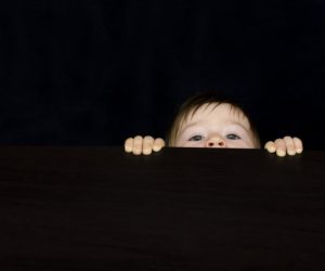 Face and hands peeking over a dark surface against a black background