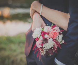 Two people embrace while one holds a bouquet behind the other