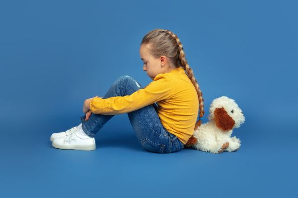 Child sitting with knees drawn up beside a stuffed dog against a blue backdrop