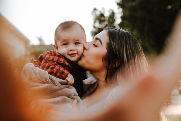 Adult holding a smiling baby and kissing the baby's cheek outdoors