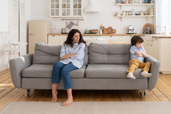 An adult and a child sit apart on a sofa with their arms crossed