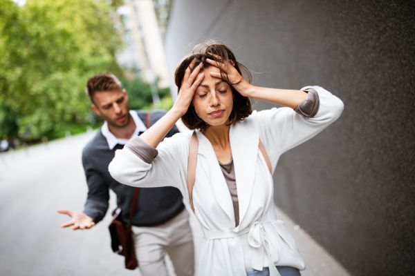 A person holds their head while another gestures behind them on a sidewalk