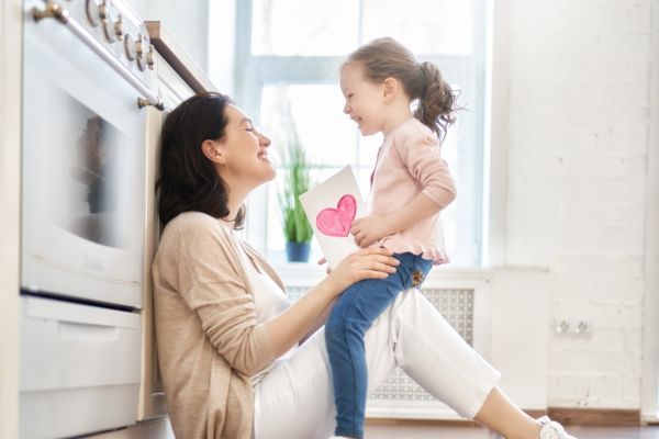 Adult smiles at a child holding a heart drawing in a bright kitchen