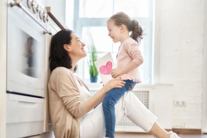 Adult smiles at a child holding a heart drawing in a bright kitchen