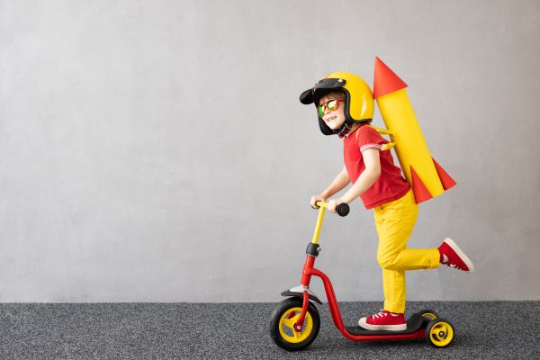 A child wearing a helmet and toy rocket backpack rides a red scooter