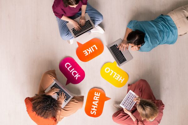 Four people use laptops around speech bubbles reading LIKE, CLICK, HELLO, and SHARE