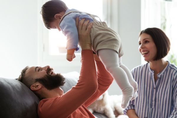 Adult lying on a sofa lifts a baby while another adult smiles nearby