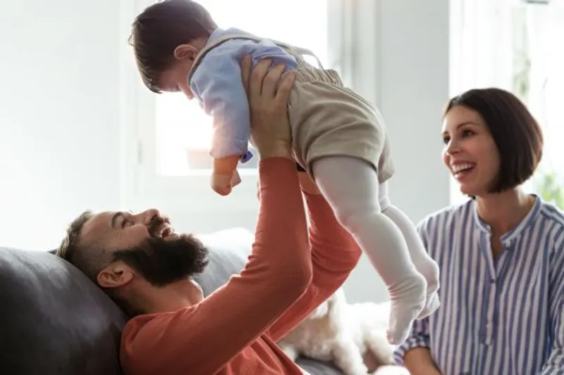 Adult lying on a sofa lifts a baby while another adult smiles nearby