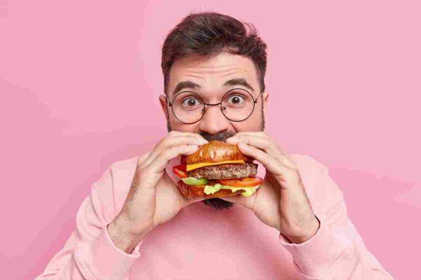 Person with glasses taking a bite from a large hamburger against a pink background