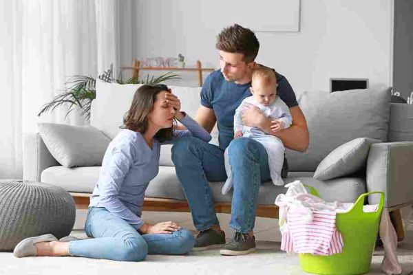 Adult holds a baby while another sits on the floor with a hand on their forehead