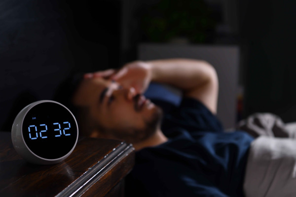 Person lies in bed beside a digital clock displaying 02:32