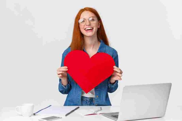 Person with glasses holding a large red heart behind a desk with a laptop