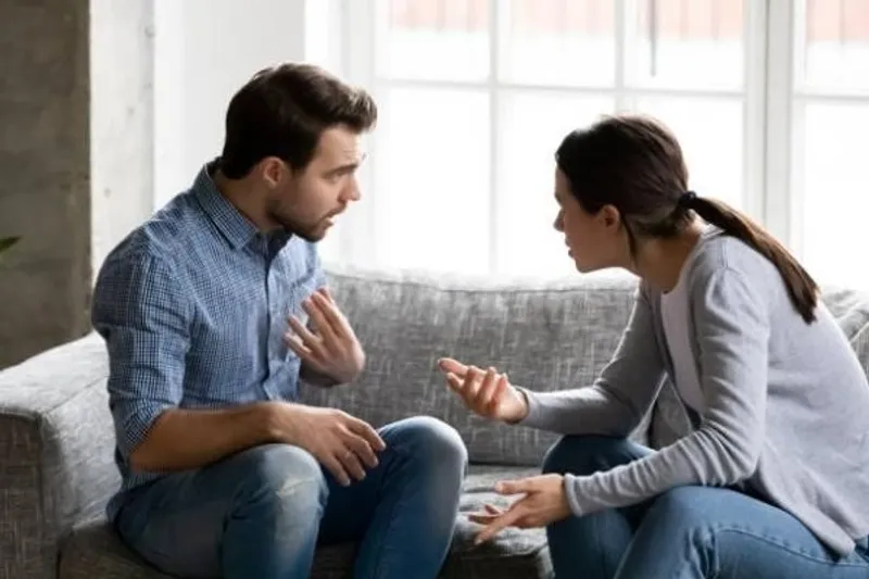 Two adults seated on a sofa, facing each other and gesturing as they talk