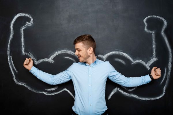 Person flexing both arms in front of chalk-drawn muscular arms on a blackboard