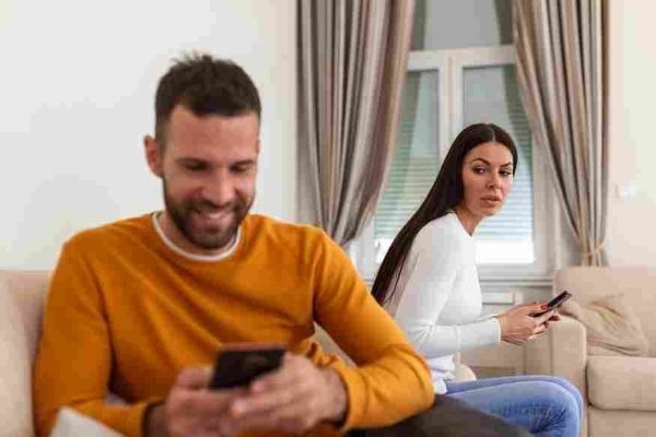 One person smiles at a phone while another looks over from a sofa