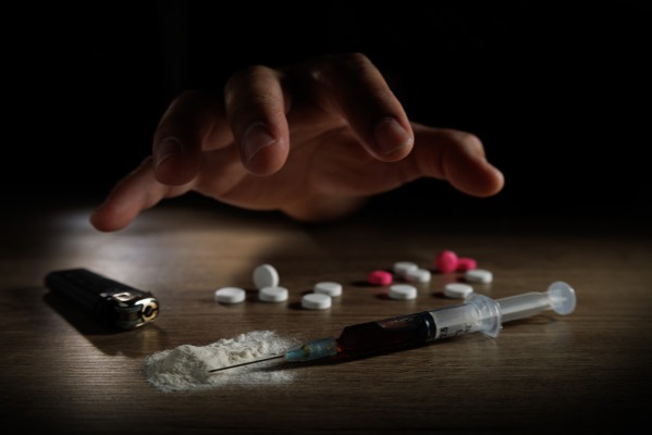 Hand hovering above a syringe, scattered pills, white powder, and a lighter