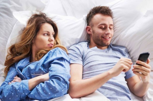 Two adults lie in bed as one watches the other use a phone