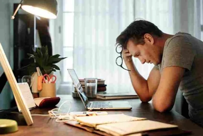 Person at a cluttered desk resting their head on one hand beside a laptop
