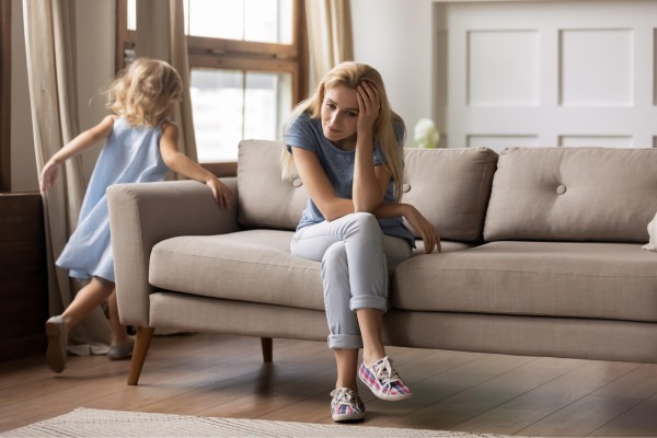Adult holding their forehead while seated on a sofa as a child moves behind them