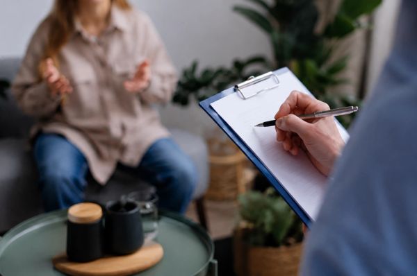 Therapist writing notes on clipboard while client speaks in a calm counseling session