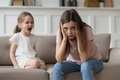 Adult covers their ears as a child sits with mouth open beside them on a sofa