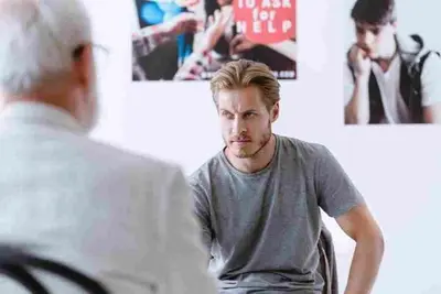 A seated person faces another person in a bright room with posters on the wall