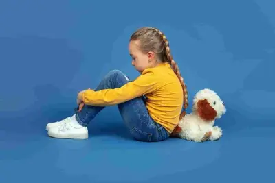 Child sitting with knees drawn up beside a stuffed dog against a plain backdrop