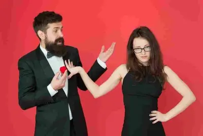 Bearded person holds out a small red box while another gestures away