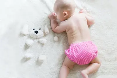 Infant lying on a white blanket in a pink diaper beside a teddy bear