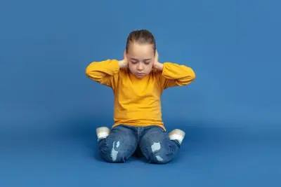 Child kneeling with both hands covering their ears against a plain backdrop