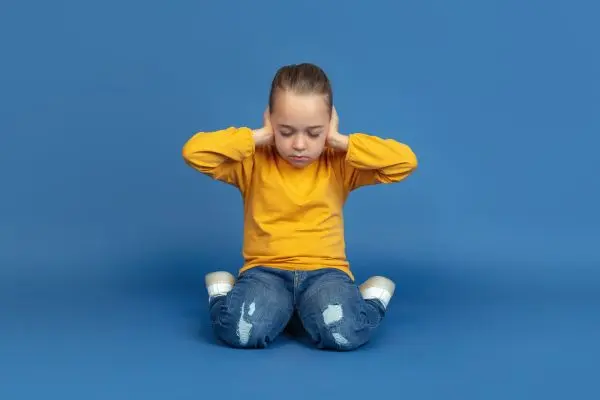 Child kneeling with both hands covering their ears against a plain backdrop