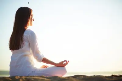 Person sits cross-legged with one hand raised beside a calm body of water