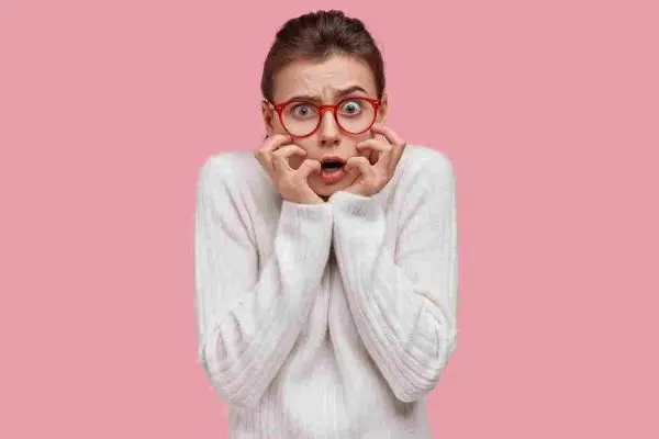 Person in red glasses holding their hands to their mouth against a pink background