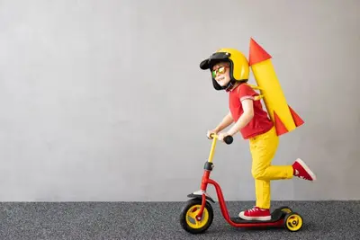 A child wearing a helmet and toy rocket backpack rides a red scooter