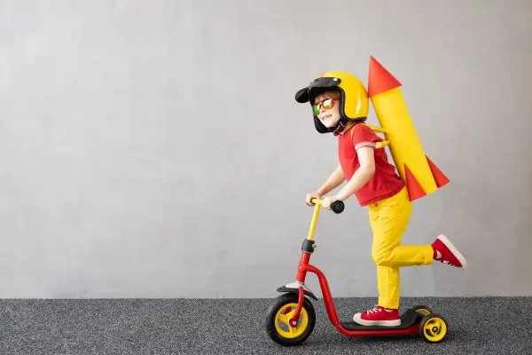 A child wearing a helmet and toy rocket backpack rides a red scooter