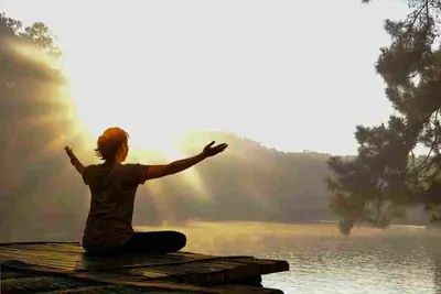 Person sitting cross-legged with arms extended beside a sunlit lake