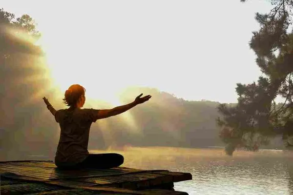 Person sitting cross-legged with arms extended beside a sunlit lake