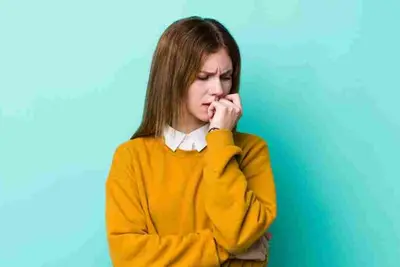 Person in a yellow sweater bites a fingernail against a plain wall