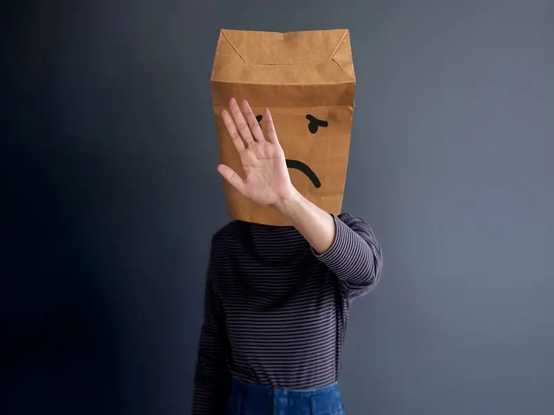 Person wearing a sad-faced paper bag and raising one hand