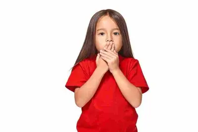 Child in a red shirt covering their mouth with both hands against a white background