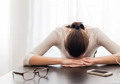 Person resting face-down on folded arms at a table with glasses and a phone