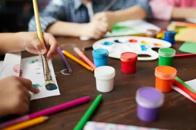 Hands painting on paper at a table covered with brushes, pencils, and paint pots