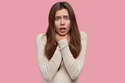 Person with wide eyes holding both hands at their throat against a pink background