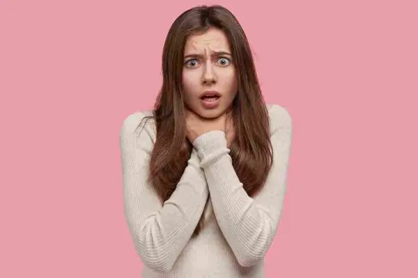 Person with wide eyes holding both hands at their throat against a pink background