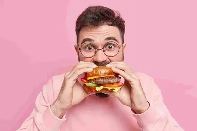 Person with glasses taking a bite from a large hamburger against a pink background