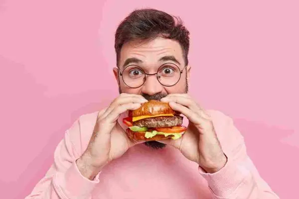 Person with glasses taking a bite from a large hamburger against a pink background