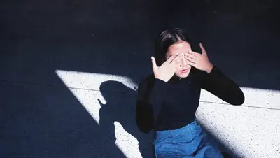 Person seated on the floor covering their eyes with both hands in a shaft of sunlight